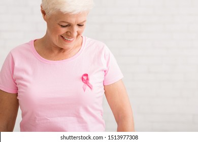 Breast Cancer. Smiling Elderly Woman Looking At Pink Ribbon On T-Shirt Standing Over White Wall Indoor. Copy Space, Shallow Depth - Powered by Shutterstock