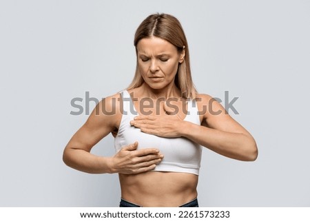 Breast screening. Anxious middle aged blonde woman wearing white top examining her breasts, grey studio background, cancer prevention concept, copy space [[stock_photo]] © 