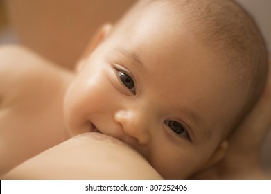 Breastfeeding baby. Smiling infant looking at her mothers eyes, while breast feeding in a cross cradle hold position. Happy Caucasian newborn looking at camera during nursing. Motherhood background.