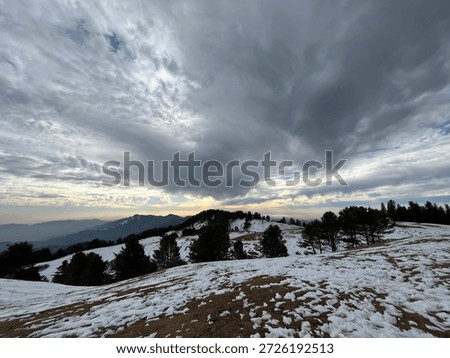Breathless panoramic view of snow-capped mountains under a dramatic cloudy sky at sunset. Beautiful winter landscape featuring rolling hills covered in white snow, pine tree forests, and a golden horizon. Scenic Himalayan mountain range view during a cold winter evening, perfect for nature and travel backgrounds