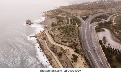 Breathtaking aerial view of coastal highway on cliffside with crashing waves, misty ocean, rocky terrain, and scattered houses. - Powered by Shutterstock