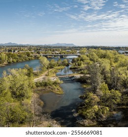 A breathtaking aerial view of lush greenery surrounding a winding river, with bridges and a distant mountain range under a vibrant, partly cloudy sky - Powered by Shutterstock