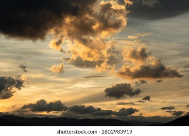 A breathtaking cloudscape captured during the height of the golden hour. Drifting, textured storm clouds are illuminated with vibrant shades of gold, amber, and orange against patches of soft evening sky, hovering peacefully over the dark, layered silhouettes of a distant mountain range at twilight.