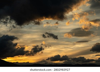 A breathtaking cloudscape captured during the height of the golden hour. Drifting, textured storm clouds are illuminated with vibrant shades of gold, amber, and orange against patches of soft evening sky, hovering peacefully over the dark, layered silhouettes of a distant mountain range at twilight.