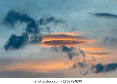 A breathtaking cloudscape captured during the height of the golden hour. Drifting, textured storm clouds are illuminated with vibrant shades of gold, amber, and orange against patches of soft evening sky, hovering peacefully over the dark, layered silhouettes of a distant mountain range at twilight.