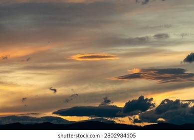 A breathtaking cloudscape captured during the height of the golden hour. Drifting, textured storm clouds are illuminated with vibrant shades of gold, amber, and orange against patches of soft evening sky, hovering peacefully over the dark, layered silhouettes of a distant mountain range at twilight.