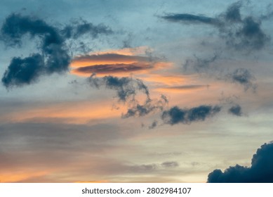 A breathtaking cloudscape captured during the height of the golden hour. Drifting, textured storm clouds are illuminated with vibrant shades of gold, amber, and orange against patches of soft evening sky, hovering peacefully over the dark, layered silhouettes of a distant mountain range at twilight.