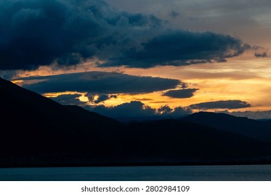 A breathtaking cloudscape captured during the height of the golden hour. Drifting, textured storm clouds are illuminated with vibrant shades of gold, amber, and orange against patches of soft evening sky, hovering peacefully over the dark, layered silhouettes of a distant mountain range at twilight.