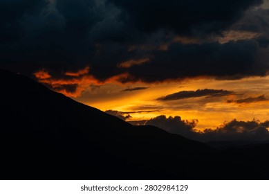 A breathtaking cloudscape captured during the height of the golden hour. Drifting, textured storm clouds are illuminated with vibrant shades of gold, amber, and orange against patches of soft evening sky, hovering peacefully over the dark, layered silhouettes of a distant mountain range at twilight.
