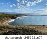 Breathtaking coastal landscape with a sandy beach, rolling green hills, and mountain backdrop under a cloud-streaked blue sky