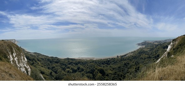 A breathtaking coastal panorama with cliffs framing lush green vegetation and a calm blue sea under a vast sky streaked with clouds, capturing the beauty of nature and expansive seaside landscapes. - Powered by Shutterstock