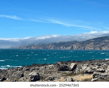 A breathtaking coastal scene with rocky shores and turquoise waters. Distant cliffs are partially covered in mist under a clear blue sky, showcasing the raw beauty of nature along the rugged coastline - Powered by Shutterstock