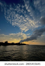 Breathtaking dramatic sky with scattered altocumulus cloud formations during golden hour sunset over a tropical island silhouette in Riau Islands, Indonesia. Colorful gradient sky from orange to deep blue with textured ocean waves in foreground.