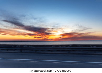 Breathtaking dramatic sunset with vibrant orange, red and yellow clouds over a calm body of water. Shot from a road or bridge with guardrail in the foreground. The sky is filled with intense colors and beautiful cloud formations during golden hour. Perfect for travel, road trip, landscape, nature and atmospheric projects.