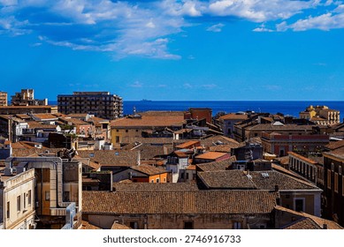 A breathtaking elevated panorama captures the historic rooftopscape of Catania, Sicily, Italy, with the deep blue expanse of the Ionian Sea stretching magnificently across the horizon under a vibrant Mediterranean sky. The dense urban fabric of terracotta-tiled rooftops fills the foreground and middle ground in a warm mosaic of russet, orange, and amber tones — satellite dishes, chimneys, air conditioning units, and rooftop terraces adding authentic lived-in detail to the scene. The buildings range from elegant ochre and coral-pink historic facades to more utilitarian mid-century apartment blocks, reflecting Catania's layered architectural history. A distant ship is faintly visible on the glittering sea horizon, reinforcing the coastal character of the city. The brilliant cobalt blue of the Ionian Sea creates a striking horizontal band of color separating the warm urban mass below from the vivid sky above, where dramatic white cumulus clouds billow against the deep blue. A lighthouse or tall column is barely discernible at the waterfront. The composition masterfully balances the dense, warm-toned city with the cool, expansive sea and sky, creating a powerful sense of place that is unmistakably Sicilian — a sun-drenched coastal city with centuries of history embedded in every rooftop.