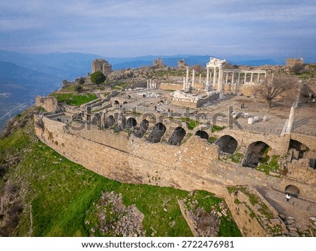 Breathtaking elevated view of Pergamon Acropolis in Turkey, showcasing the iconic white marble Temple of Trajan with dramatic columns, vast ancient theater terraces, robust stone walls with intricate arches framed by lush green grass, distant rugged mountains, and a vibrant partly cloudy sky at golden hour. Tiny tourists explore this UNESCO World Heritage site, perfect for travel ads, history visuals, and epic landscapes.