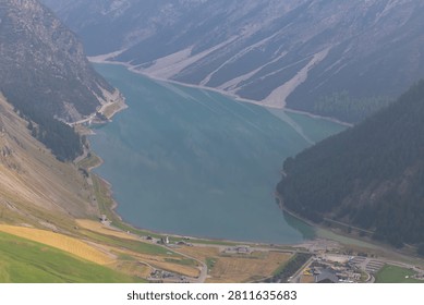 Breathtaking high angle view of a long turquoise alpine lake nestled between deep valleys, pine forests and steep mountain ridges on a hazy day.