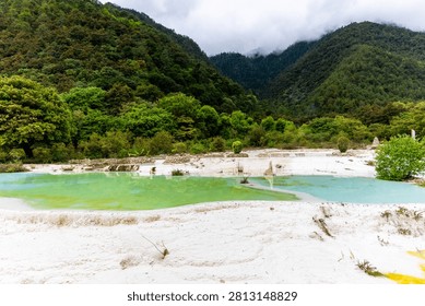 A breathtaking high dynamic range (HDR) landscape of the Baishuitai travertine terraces in Yunnan. The image showcases shallow,natural pools filled with vibrant green and light blue mineral water,bordered by the bright white calcium carbonate deposits. Lush green trees and shrubs grow directly out of the geological formations. In the background,majestic,densely forested mountains rise sharply,their peaks touching a moody,low-hanging cloud cover,highlighting the raw and wild beauty of this remote mountain region.