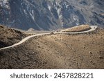 A breathtaking high-altitude road snakes through rugged mountains under a blue sky, with distant snow-covered peaks.