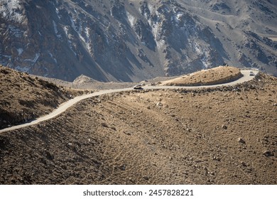 A breathtaking high-altitude road snakes through rugged mountains under a blue sky, with distant snow-covered peaks. - Powered by Shutterstock