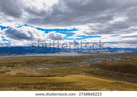 A breathtaking high-angle view overlooking the town of Litang and the vast meadow plains. In the distance, the majestic snow-capped mountains of the Shaluli Range stand against a cloudy sky, defining the horizon of the 