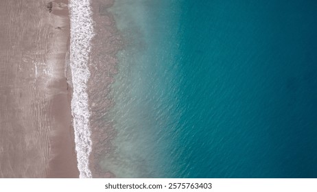 Breathtaking high-angle view of a sandy beach meeting a deep teal ocean. Waves break gently on the shore, creating a stunning contrast that captures the essence of coastal paradise