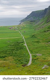 A breathtaking high-angle view from a summit overlooking Unstad village in Norway's Lofoten Islands, where green fields meet dramatic cliffs and the vast Arctic Ocean under a moody summer sky.
This high-angle view highlights a dramatic coastal landscape where a lush valley meets the Norwegian Sea.

Unstad Beach is world-famous as a premier Arctic cold-water surfing destination. It is recognized as one of the northernmost surf spots in the world, attracting professional and adventurous surfers globally due to its highly consistent swell.

Lush Fields: Bright green, rolling meadows cover the entire valley floor.
Steep Ridges: Rocky, grass-covered mountains rise sharply on the right side.
Coastal Incline: The land slopes gently downward toward a crescent-shaped bay.

The Ocean: A calm, expansive body of grey-blue water fills the background.
The Shoreline: A distinct coastal border separates the green fields from the sea.
Overcast Sky: Muted, grey clouds diffuse the daylight evenly across the landscape.