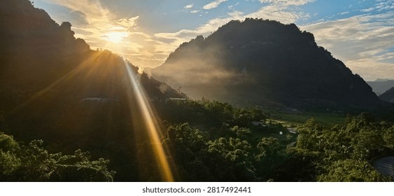 A breathtaking landscape shot capturing a dramatic golden sunset over the mountains in Thổ Bình commune, Tuyên Quang province, Vietnam. A powerful sunbeam cuts diagonally across the foreground, while a layer of evening mist and fog settles in the lush, green tropical valley below the steep mountain ridges under a warm twilight sky