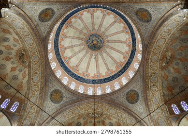 Breathtaking low-angle view looking directly up at the massive central dome of a historic mosque, adorned with intricate blue and red geometric patterns, arabic calligraphy, and circular medallions. Sunlight filters through stained glass windows, illuminating the detailed frescoes and architectural grandeur.