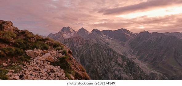 A breathtaking mountain range at sunset with dramatic clouds, rocky ridges, and lush green slopes. Captured from a high-altitude trail with a golden light casting a dreamy atmosphere.