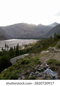 A breathtaking panoramic high-angle landscape view overlooking a lush green mountain valley village, terraced fields, and rural houses. A wide, winding river flows through the valley floor below, surrounded by majestic, rugged mountains under a clear sky. In the foreground, a sloping hillside features wild grass, rocks, and an earthy trail. This high-resolution travel and nature stock photograph highlights rural life, scenic geography, eco-tourism, outdoor adventures, and remote settlements in a stunning mountainous region, making it ideal for backgrounds, tourism marketing, environmental projects, and editorial concepts.