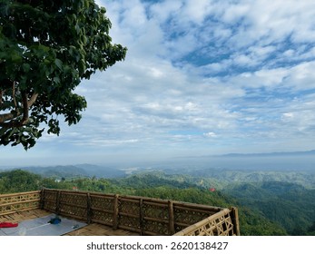 A breathtaking panoramic view from a bamboo deck overlooking the lush green hills of Bandarban, Bangladesh. The vibrant landscape stretches under a sky filled with scattered clouds.