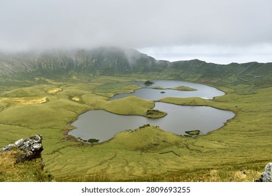 A breathtaking panoramic view of the caldera with its tranquil lakes, surrounded by lush green terraced slopes and dramatic rock formations under a cloudy sky, Corvo Island, Azores.