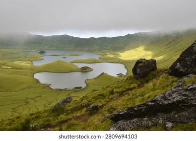 A breathtaking panoramic view of the caldera with its tranquil lakes, surrounded by lush green terraced slopes and dramatic rock formations under a cloudy sky, Corvo Island, Azores.
