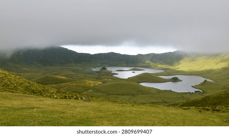 A breathtaking panoramic view of the caldera with its tranquil lakes, surrounded by lush green terraced slopes and dramatic rock formations under a cloudy sky, Corvo Island, Azores.