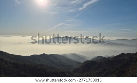 A breathtaking panoramic view captured from the mid-slopes of Gwanaksan Mountain near Seoul, South Korea. A dense layer of morning fog blankets the city below, creating a dramatic sea of clouds. The ridgeline of Cheonggyesan Mountain emerges clearly above the mist, while rolling green hills frame the foreground. The sun shines brightly against a vivid blue sky, casting a serene and otherworldly atmosphere over the layered mountain silhouettes.