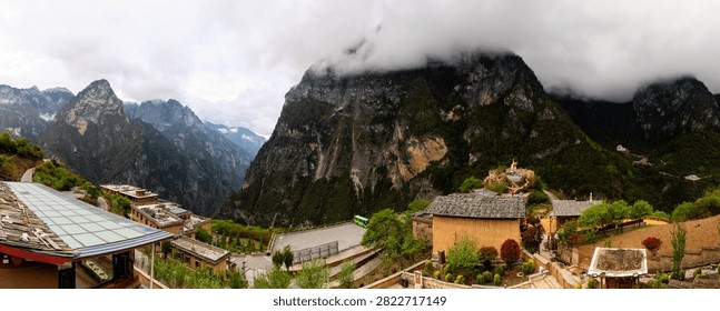 A breathtaking panoramic view of a deep mountain gorge and a traditional settlement in Yunnan, China. The vast landscape features towering rock faces, heavy, low-hanging clouds, a winding mountain road, and a hillside Buddhist shrine adorned with colorful prayer flags.