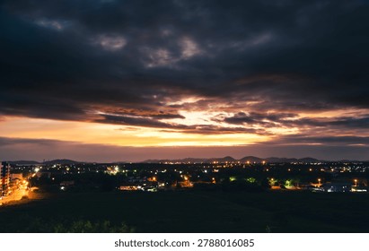 A breathtaking panoramic view of a dramatic sunset over a suburban town. The sky is filled with dark, moody clouds, sharply contrasted by a vibrant strip of glowing orange and yellow sunlight near the horizon. The landscape below features warm, illuminated city street lights and silhouettes of distant mountains. Perfect for concepts of twilight, weather changes, and tranquil evenings.