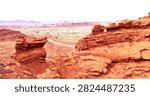 Breathtaking panoramic view of Utah’s iconic red rock desert, where layered rust-colored sandstone formations and whimsical hoodoo spires frame a vast valley stretching to hazy distant mesas. A winding dirt road cuts through sun-baked earth dotted with hardy desert shrubs, while ancient wind-sculpted cliffs rise in rich terracotta hues under a bright pale sky. This timeless scene captures the raw geological beauty, immense scale, and legendary wild grandeur of the American Southwest’s Canyon Country.