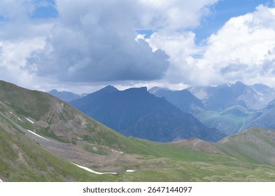 Breathtaking panoramic view of a mountain range with lush green alpine meadows in the foreground, dramatic blue peaks in the distance, patches of snow, and a cloudy blue sky overhead.