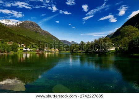 A breathtaking panoramic view of a tranquil river or lake in the St Stryn region of Norway, with crystal-clear water reflecting the vibrant blue sky and scattered white clouds. Lush green mountains rise majestically on both sides, with patches of snow visible on their peaks. A charming village featuring traditional Norwegian houses is nestled along the waterfront and on the lower slopes, surrounded by dense green trees and foliage. The scene perfectly captures the pristine natural beauty and peaceful atmosphere of the Scandinavian landscape.
