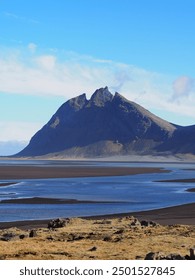 A breathtaking, rugged mountain rises dramatically above the calm water below, set against a clear blue sky, providing a stunning natural view and sense of adventure - Iceland.