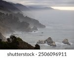 A breathtaking and serene view of the rugged, dramatic coastline of Big Sur, located in California, where mist softly envelops the lofty cliffs and gentle waves lap peacefully at the shore