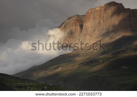 Breathtaking summer mountain landscape - high orange rocky cliff of canyon in in golden sunbeam and shadows flowing fluffy cloud on green velvety hilly slope, grey stormy sky. Majestic panorama view.