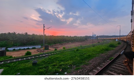 A breathtaking sunset captured from a moving train, showcasing vibrant orange and pink hues across the sky. The railway tracks curve through lush green vegetation and open land, creating a dynamic perspective. Overhead electric lines and distant trees add depth, while the peaceful countryside atmosphere highlights the beauty of travel and nature.