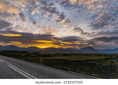 A breathtaking sunset landscape featuring a dramatic sky filled with layered clouds illuminated in golden orange and deep purple hues. A paved road with white lane markings curves through the foreground, leading the eye toward a vast volcanic plain with sparse green moss vegetation. Snow-capped mountains create a striking silhouette along the horizon as the sun sets behind them, casting warm golden light through breaks in the clouds. The scene evokes Iceland's rugged wilderness and the magic of midnight sun conditions.