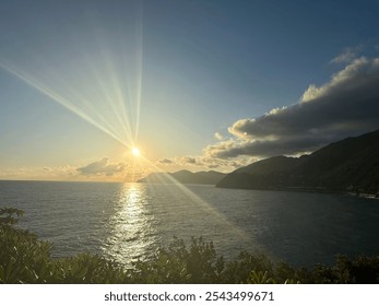 A breathtaking sunset over a serene sea, casting golden reflections on the water. Silhouetted mountains and dramatic clouds frame the horizon, while sun rays burst through, illuminating the scene. - Powered by Shutterstock