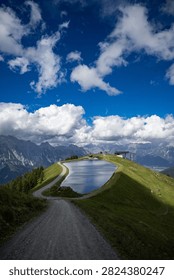 Breathtaking vertical panoramic view of the alpine reservoir lake captured from the absolute summit of Großer Asitz in Leogang, Austria. In the foreground, a wide gravel hiking path made of small grey stones and pebbles curves beautifully down the ridge, leading the eye towards a crystal-clear artificial pond. The calm water surface perfectly reflects the dramatic cloudy sky, nestled on a vibrant green grassy hill that is striking divided by a sharp natural cloud shadow. In the midground, the scenic walking track encircles the lake, leading to a modern ski resort cable car station on the horizon. In the far background, the spectacular, sharp rocky peaks and grey stone formations of the majestic Leogang Mountains rise grandly under an expansive, deep blue summer sky filled with massive, fluffy white cumulus clouds. This stunning scenery perfectly captures the peaceful essence of alpine eco-tourism, mountain infrastructure, and outdoor trekking adventure in the Austrian Alps. Ideal for vertical mobile backgrounds, travel agency banners, and nature-themed commercial advertising projects.