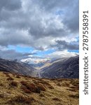 A breathtaking vertical view looking down a deep alpine valley with a winding river and road, surrounded by autumn mountain slopes under thick dramatic clouds.