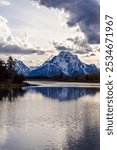 Breathtaking view of the Grand Teton mountain range, beautifully reflected in a calm lake in Wyoming. Dramatic clouds enhance the peaceful and majestic landscape, offering a serene natural scene.